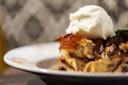 A close-up of a dessert featuring bread pudding topped with a scoop of vanilla ice cream and caramel shards, served on a white plate. The background is softly blurred.