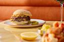 A cheeseburger with a sesame seed bun and chopped toppings sits on a white plate with two green chili peppers, next to a glass of red wine and a lemon wedge on a glossy table.