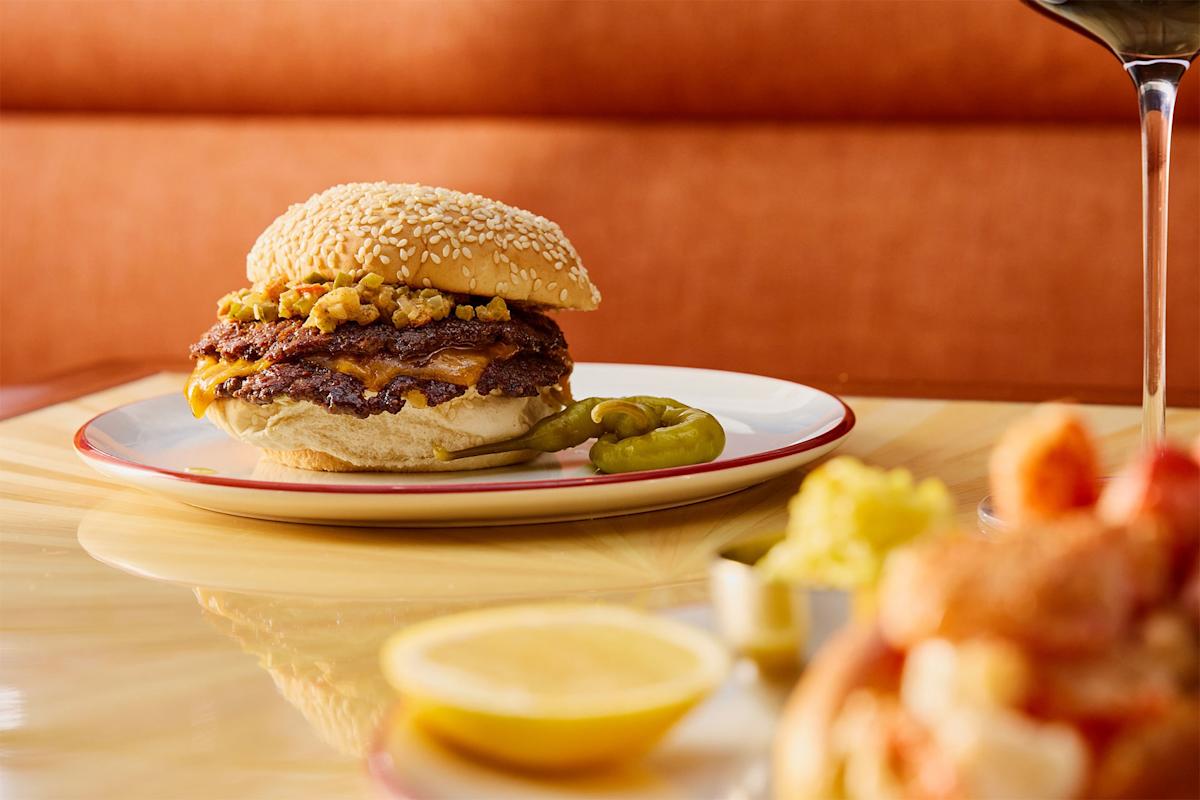 A cheeseburger with a sesame seed bun and chopped toppings sits on a white plate with two green chili peppers, next to a glass of red wine and a lemon wedge on a glossy table.