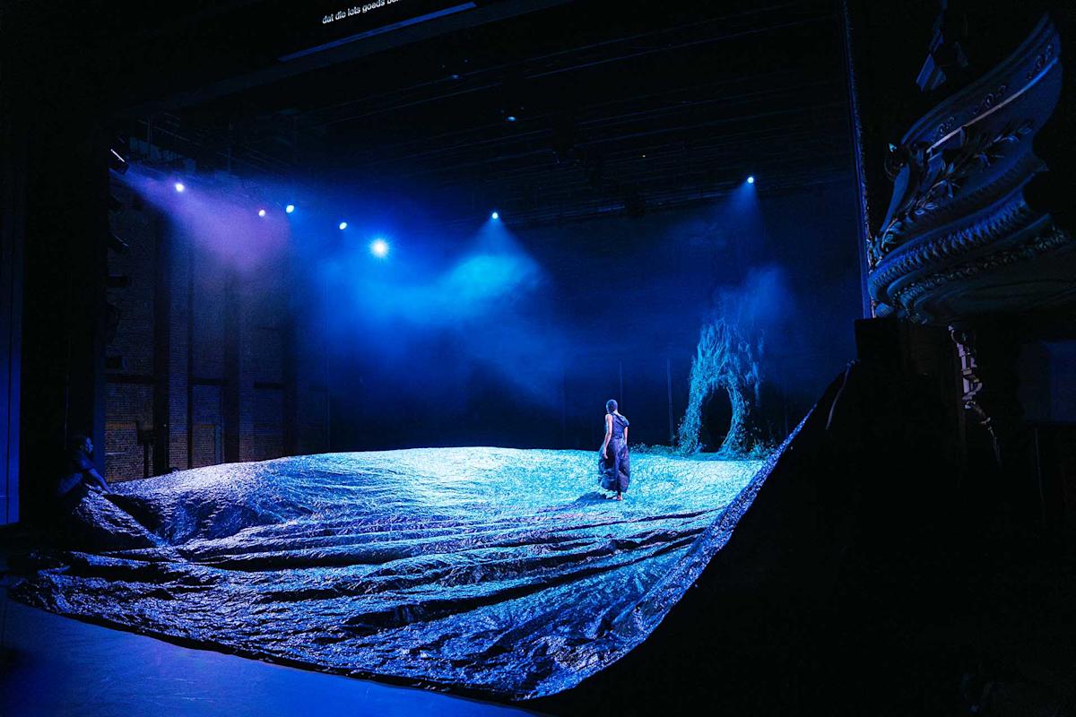 A person stands on a stage covered in textured blue material, resembling waves, under dramatic blue lighting with mist and a backdrop suggesting a cave or ocean scene.