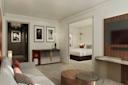 Modern hotel suite with a living area featuring a sofa, armchair, coffee table, and wall art; a TV mounted on the wall; and a view into a bedroom with a neatly made bed. Neutral tones and stylish decor throughout.