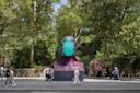 Several people walk past a large, colorful sculpture of a head wearing a turquoise face mask, set on a city sidewalk surrounded by tall trees and greenery.