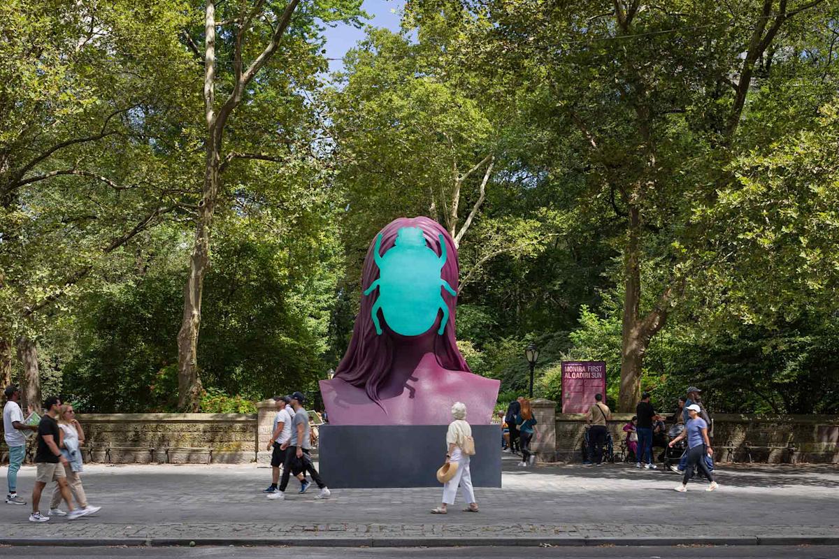 Several people walk past a large, colorful sculpture of a head wearing a turquoise face mask, set on a city sidewalk surrounded by tall trees and greenery.