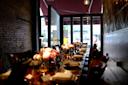 A long wooden dining table is set for a meal with plates, glasses, and small floral arrangements. The table is in a dimly lit restaurant with exposed brick walls and large windows letting in natural light from a partially open door.