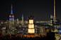 A night view of New York City featuring the illuminated Empire State Building and surrounding skyscrapers. In the foreground, people stand on a brightly lit tiered platform observing the cityscape.