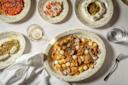 A table set with plates of seafood pasta, shrimp, creamy cheese with herbs, a dish with feta and seeds, a glass of white wine, and a glass of water on a white tablecloth.