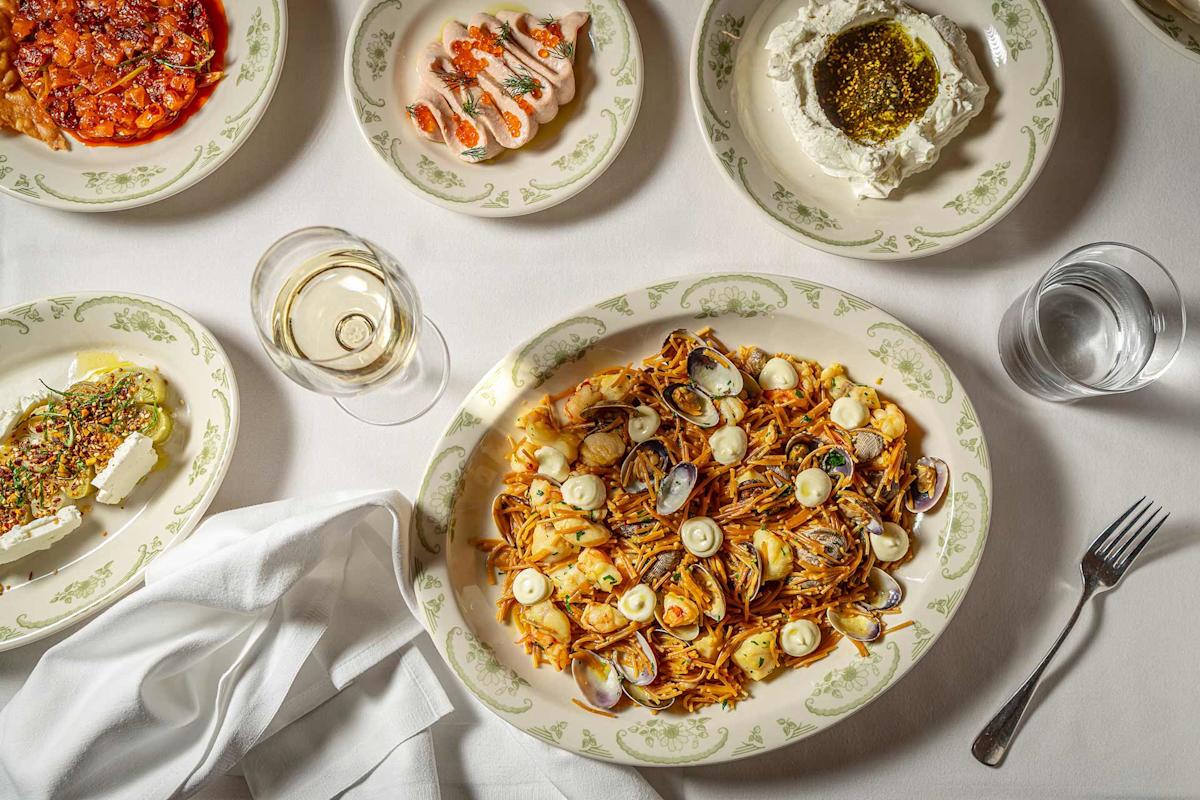 A table set with plates of seafood pasta, shrimp, creamy cheese with herbs, a dish with feta and seeds, a glass of white wine, and a glass of water on a white tablecloth.