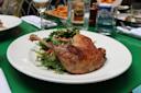 A plate with a roasted duck leg served on a bed of mixed greens and vegetables, set on a green table with glasses of water, wine, fries, and other dishes in the background.