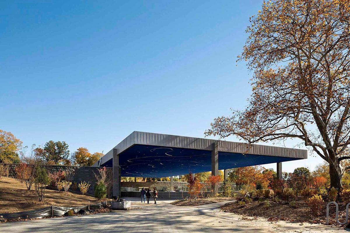 A modern pavilion with a wide, blue star-patterned ceiling stands in a park. Autumn trees with sparse leaves surround the structure. Three people walk nearby under a clear blue sky.