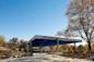 A modern pavilion with a wide, blue star-patterned ceiling stands in a park. Autumn trees with sparse leaves surround the structure. Three people walk nearby under a clear blue sky.