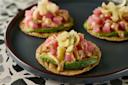 Three small tostadas on a dark plate, each topped with sliced avocado, diced raw tuna, creamy sauce, and crispy garnishes, set on a terrazzo surface.