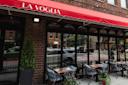 Outdoor seating at a restaurant called "La Voglia," with tables and chairs set up on the sidewalk under a red canopy, next to large windows on a city street.