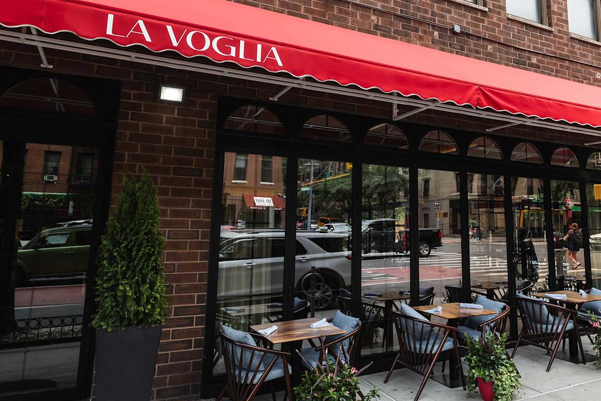 Outdoor seating at a restaurant called "La Voglia," with tables and chairs set up on the sidewalk under a red canopy, next to large windows on a city street.