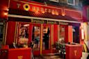 A brightly lit restaurant with red and yellow decor named Tournesol. The entrance has a red awning with “Brunch • Lunch • Dinner” written on it. Outdoor seating with green chairs and set tables is visible.