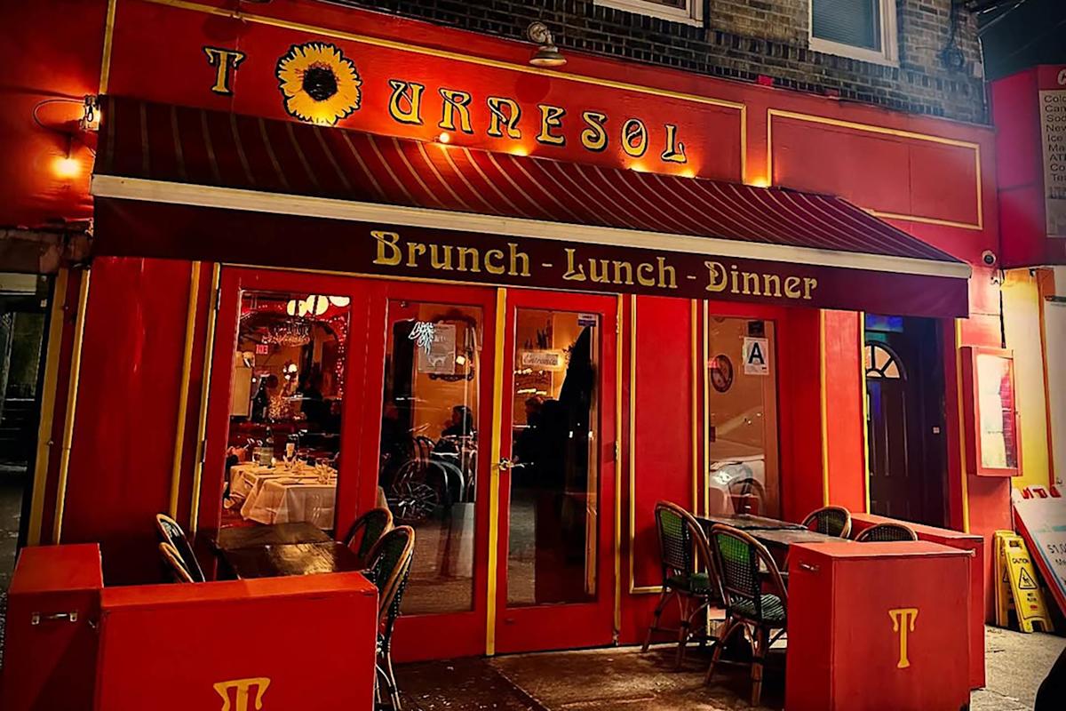 A brightly lit restaurant with red and yellow decor named Tournesol. The entrance has a red awning with “Brunch • Lunch • Dinner” written on it. Outdoor seating with green chairs and set tables is visible.