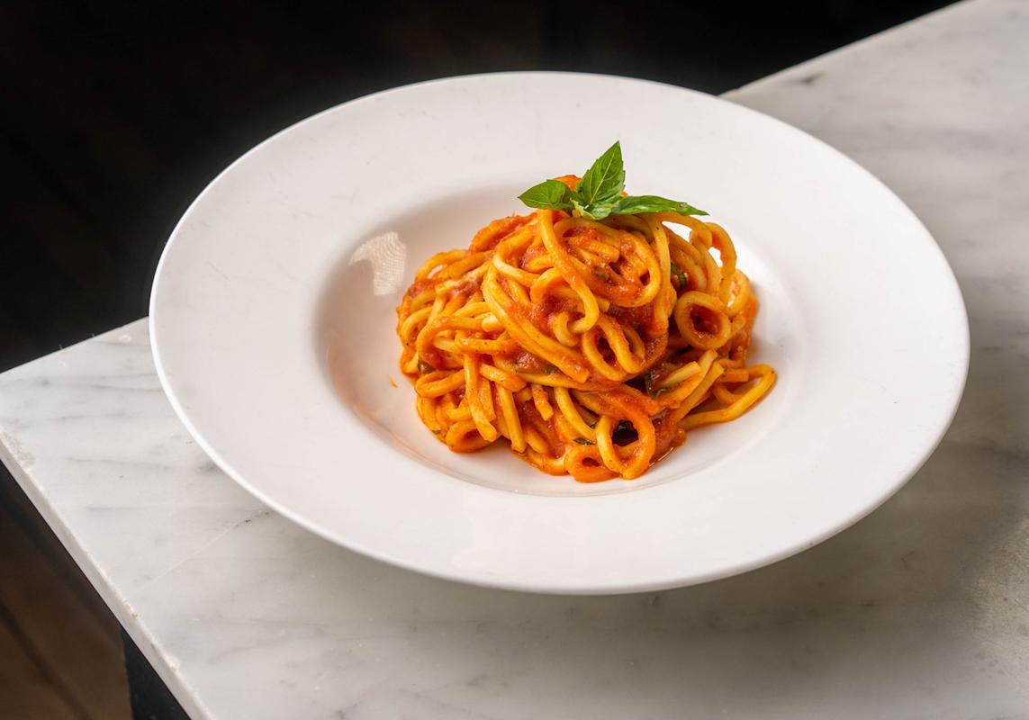 A white plate with a serving of thick spaghetti coated in tomato sauce, garnished with a sprig of fresh basil, sits on a white marble surface.