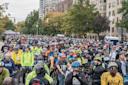 A large crowd of cyclists wearing helmets and colorful gear gathers on a city street lined with trees and tall buildings, preparing for a group ride or cycling event.