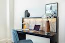 A modern desk setup with a closed laptop, a notepad, a vase of flowers, a decorative black sculpture, and a large lamp on a dark wooden desk. A soft blue chair and framed art are also visible.