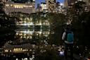 A person with a blue backpack stands by a tranquil lake at night, with city lights reflecting on the water. Trees line the opposite bank, and tall buildings illuminate the skyline in the background.