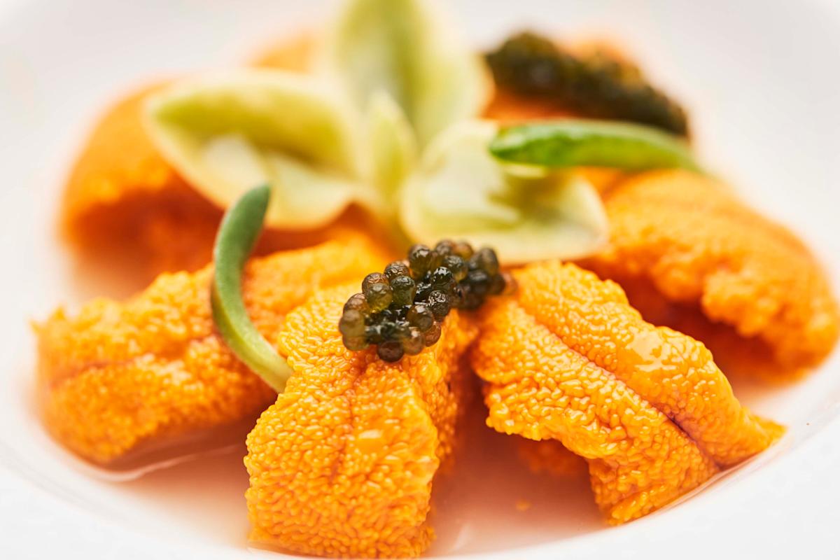 Close-up of fresh orange sea urchin (uni) topped with black caviar and garnished with green vegetables, artistically plated on a white dish.