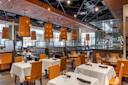 Modern restaurant interior with a mix of table and booth seating. White tablecloths, glassware, and folded napkins adorn the tables. Large cylindrical pendant lights hang from the ceiling, and a bar area is visible in the background.