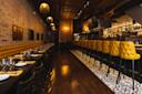 A modern restaurant interior with exposed brick walls, yellow tufted bar stools, dark wooden floors, pendant lighting, and a long bar with shelves of bottles and glassware. Tables are set and ready for guests.