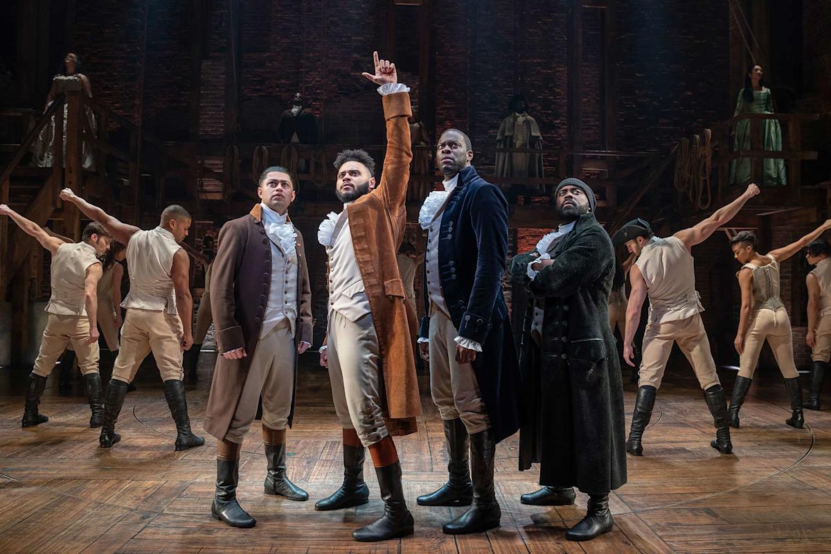 Four actors in historical costumes stand at the front of a stage, one raising his hand upward. Dancers in beige outfits pose in the background. The set resembles a wooden structure, suggesting a theatrical performance.