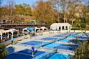 Outdoor pickleball courts in a park setting, with several people playing on different courts. Spectators watch from shaded seating areas nearby, surrounded by trees and buildings in the background.