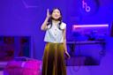 A woman in a white sleeveless top and pleated yellow skirt stands on a brightly lit stage, gesturing with her right hand. The set features purple lighting, a fuzzy pink chair, and electronics in the background.
