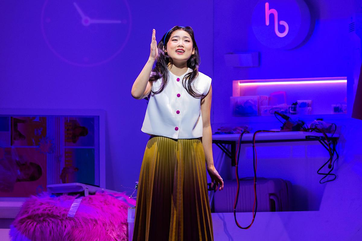 A woman in a white sleeveless top and pleated yellow skirt stands on a brightly lit stage, gesturing with her right hand. The set features purple lighting, a fuzzy pink chair, and electronics in the background.