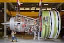 A person in safety gear inspects a large, exposed jet engine suspended in an industrial facility. The engine’s complex inner components and wiring are visible, and the person appears small beside the machinery.