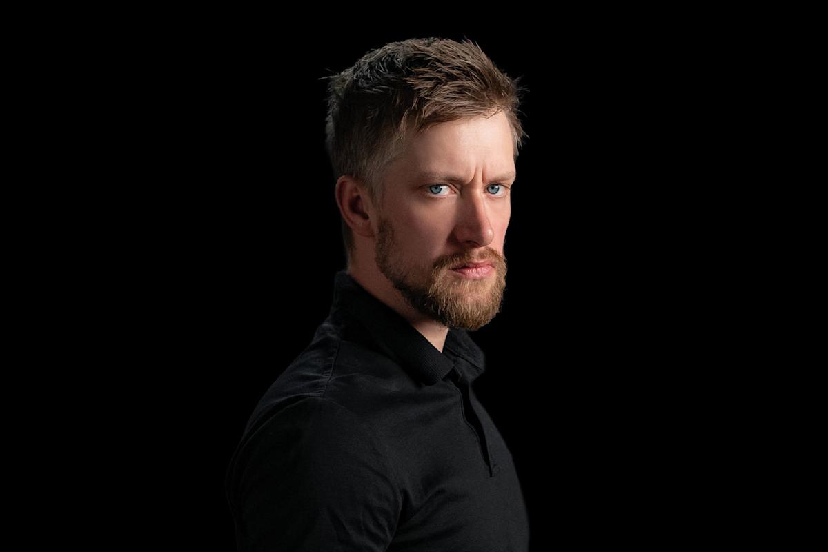 A man with short brown hair and a beard, wearing a black shirt, looks intensely at the camera against a solid black background.