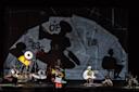 A surreal stage scene features performers in abstract, colorful costumes posed against a backdrop of newspaper clippings and bold shadows, creating an avant-garde, theatrical atmosphere.
