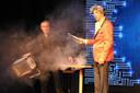 Two magicians perform on stage; one in a black suit holding a large box emitting smoke, the other in a red jacket holding metal tongs, with a digital blue background behind them.