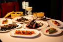 A table set with various plates of gourmet food, including lamb chops, sushi, and assorted appetizers. Two elegant cocktails are placed among the dishes. The setting is dimly lit, creating a sophisticated dining atmosphere.