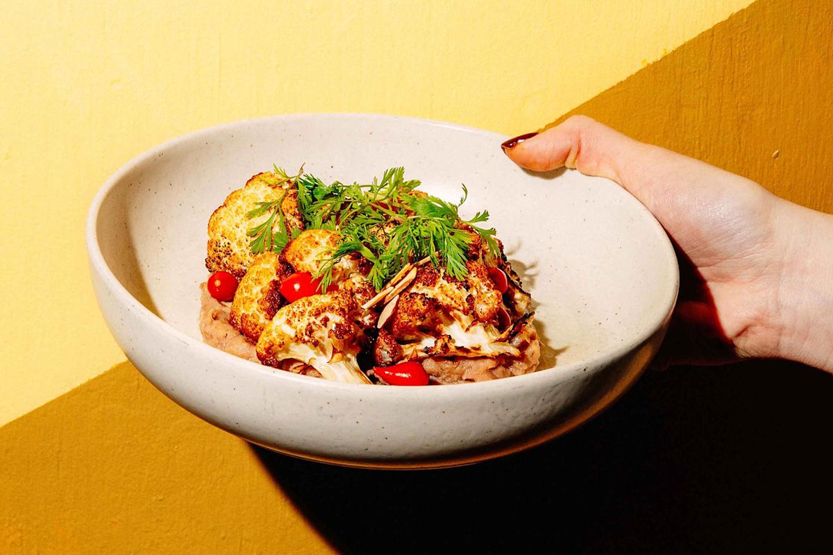 A hand holds a white bowl filled with roasted cauliflower, garnished with fresh herbs and small red peppers, against a yellow background.