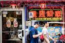 Two people walk past a colorful bakery storefront decorated with neon signs, Chinese characters, and festive paper ornaments. Bright menu images and a "Cash Only" sign are visible in the window.