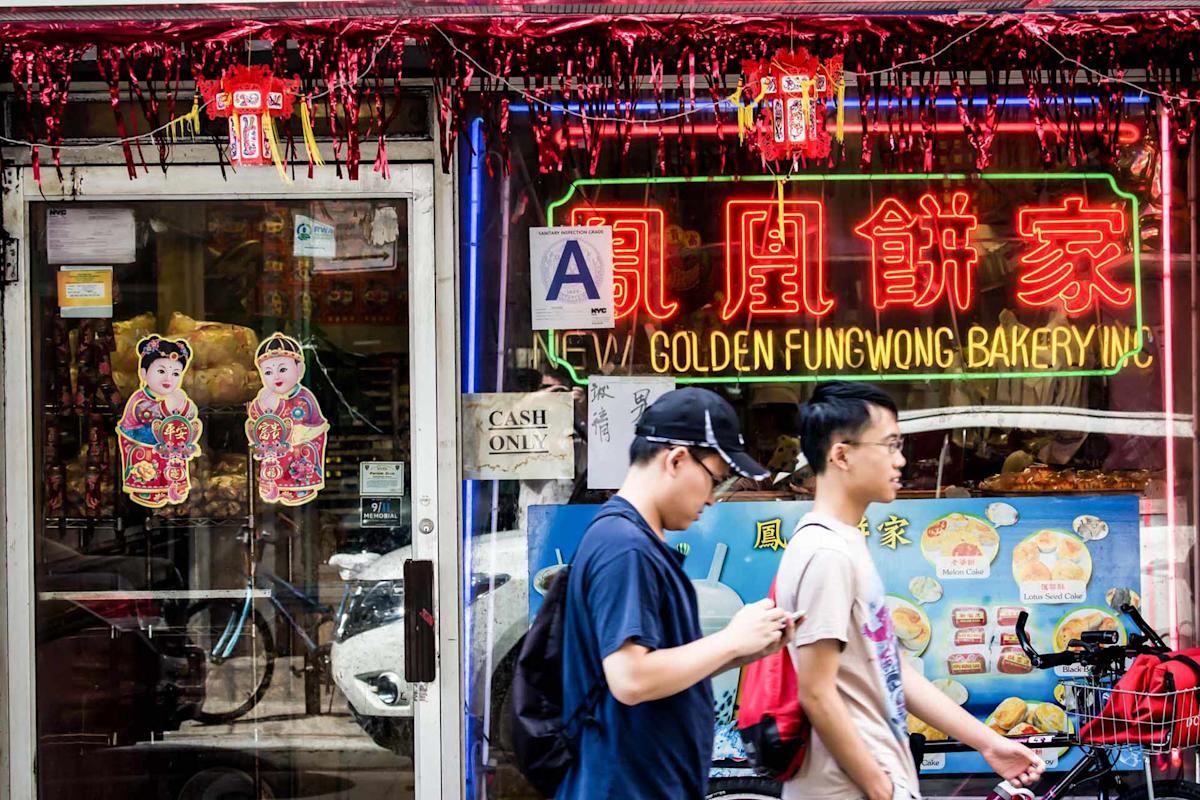 Two people walk past a colorful bakery storefront decorated with neon signs, Chinese characters, and festive paper ornaments. Bright menu images and a "Cash Only" sign are visible in the window.