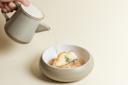 A hand pours liquid from a ceramic teapot into a round bowl containing a piece of cooked fish, garnished with herbs, on a light, neutral background.