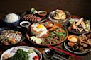 A variety of Asian dishes are arranged on a wooden table. There's rice with a fried egg, grilled meat, leafy greens, fried fish, and dipping sauces. Red chopsticks and a fork are placed nearby.