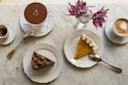 A marble table with a cappuccino and an espresso, two spoons, and three desserts: tiramisu, a chocolate cake slice, and a mango cheesecake slice. Two small vases hold loose purple flowers.