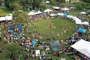Aerial view of a cultural festival with people in traditional attire dancing in a large grassy circle. Surrounding the area are white tents, trees, and spectators sitting on blankets.