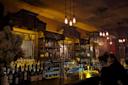 A dimly lit bar with two patrons sitting at the counter. The bar is stocked with various bottles, and vintage-style pendant lights hang from the ceiling, casting a warm glow. The atmosphere is cozy and intimate.
