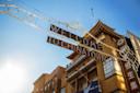 A sign reading "Welcome to Chinatown" hangs above a street with traditional Chinese-style architecture beneath a bright blue sky and sunlight.