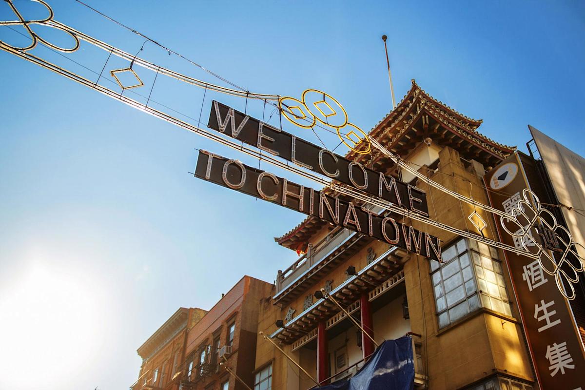 A sign reading "Welcome to Chinatown" hangs above a street with traditional Chinese-style architecture beneath a bright blue sky and sunlight.