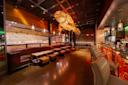 Modern restaurant interior with a large, illuminated fish-shaped light fixture, cushioned seating, a long bar with stools, and warm ambient lighting. The space features wood accents and a contemporary design.