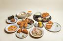 An assortment of Japanese dishes on plates, including fried cutlets, shrimp tempura, rice bowls, pickled vegetables, corn, dipping sauces, sushi, and dessert, all arranged neatly on a light background.