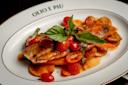 A plated dish featuring a cooked fish fillet topped with cherry tomatoes, fresh basil, and sauce, served over sliced potatoes on a white plate with "Olio e Più" written on the rim.