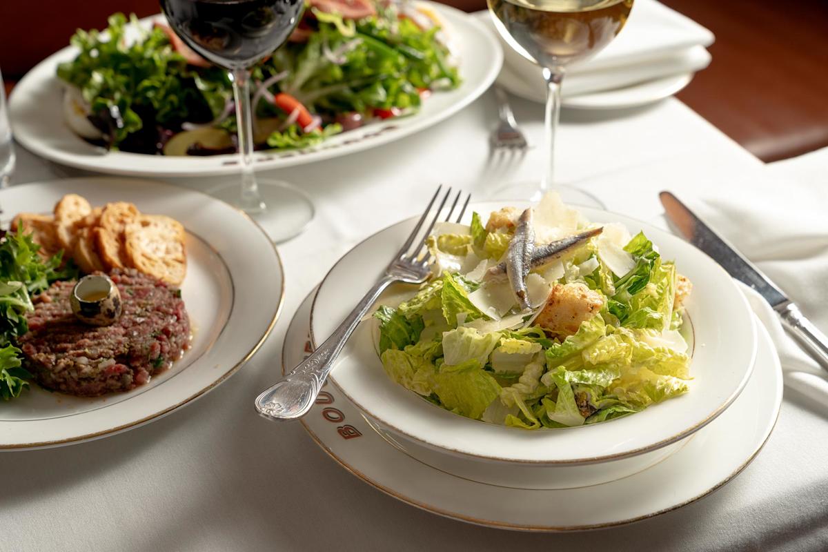 A plate of Caesar salad with lettuce, croutons, anchovies, and shaved cheese sits on a white tablecloth next to a glass of white wine, a dish of crackers, and another plate of mixed salad.