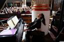 A woman passionately plays a grand piano in a large room with an audience watching. She is wearing a black outfit, and sheet music is open on the piano. The setting is elegant, with tall columns and detailed architecture.